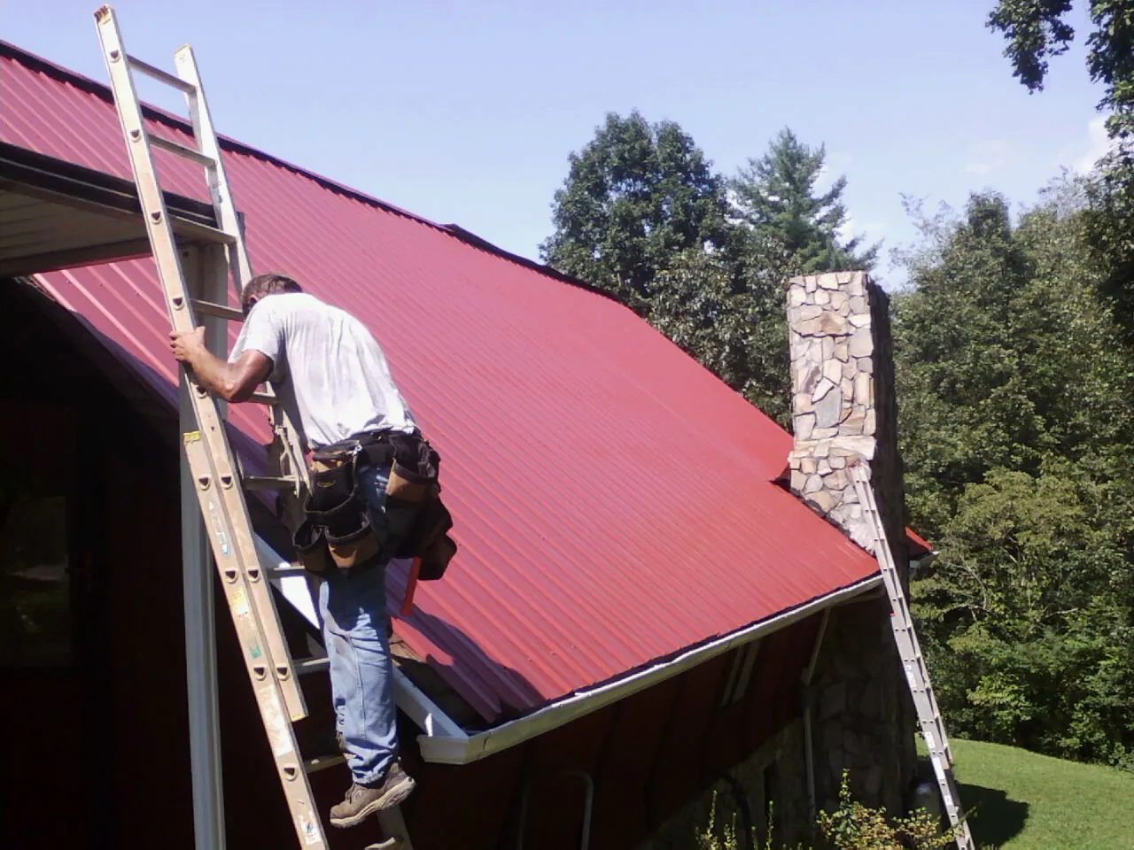 Roofer on a ladder beside a red metal roof and stone chimney — Soffit & Fascia Repair by Roofing Brothers in Fortville